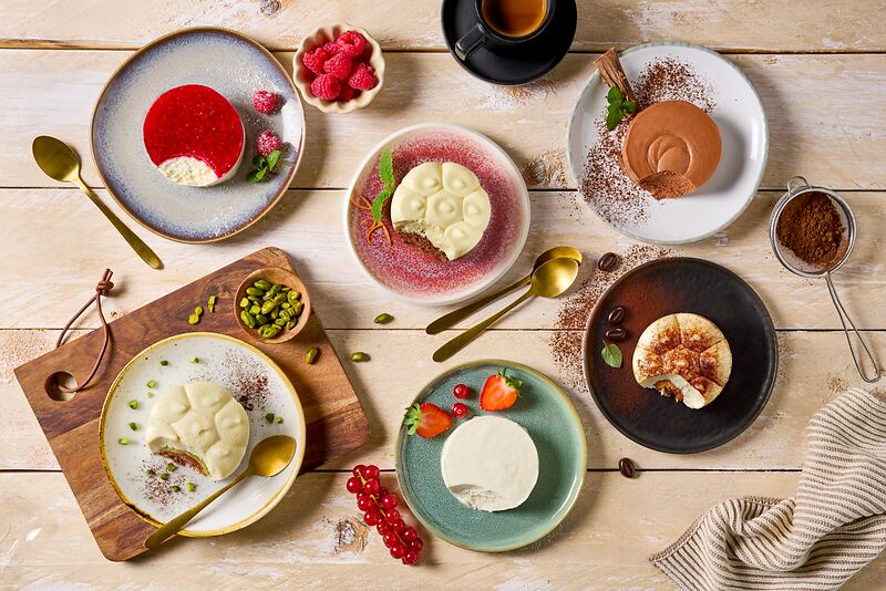 Sechs Teller mit verschiedenen Desserts, darunter Kuchen mit Beeren, Mousse und Tiramisu, stehen auf einem Holztisch, auf dem Löffel, Kaffee, Pistazien und frisches Obst verstreut sind.