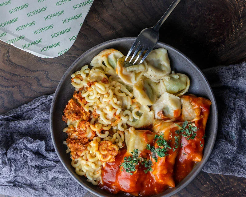 Schüssel mit verschiedenen Pastasorten: Makkaroni mit Fleischsauce, Spinat-Tortellini und Käse-Ravioli mit Tomatensauce und Petersilie. Eine Gabel darauf. Im Hintergrund eine graue Serviette und Markenpapier.