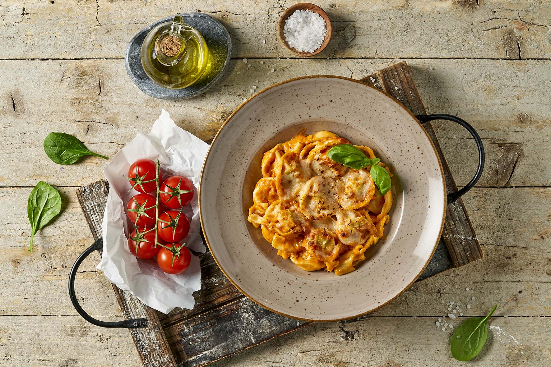 Eine Schüssel cremige Pasta mit Tomaten, Spinat, Salz und Olivenöl steht verlockend neben einem Teller mit Sattmacher der Extraklasse Fleischpflanzerl von Hofmanns Shop auf einem rustikalen Holztisch.