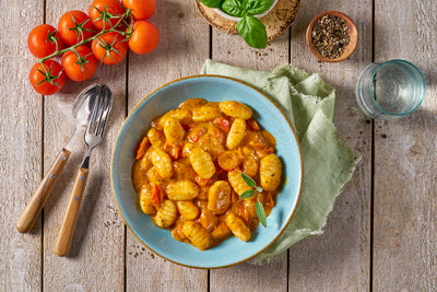 Teller mit Gnocchi mit Tomatensauce und Garnitur, daneben frische Tomaten, Basilikum, Besteck und ein Glas Wasser.
