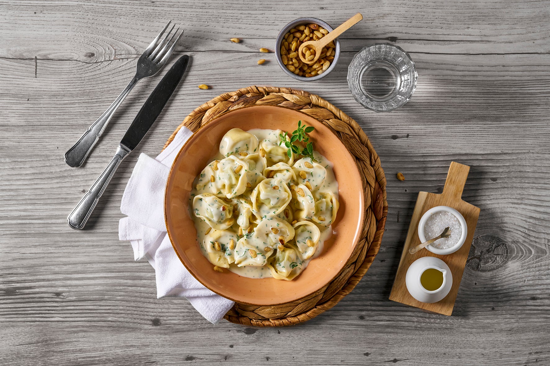 Eine Schüssel Tortelloni alla Panna von Hofmanns Shop, mit einem Glas Wasser, Besteck und Gewürzen auf einem Holztisch.