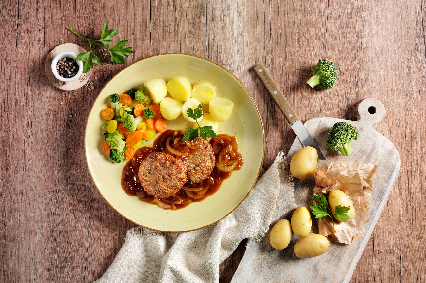 Ein Teller Sattmacher der Extraklasse aus Hofmanns Shop, bestehend aus Hackbraten mit Soße, Kartoffeln und gemischtem Gemüse, fängt die herzhafte Essenz ein, die an Fleischpflanzerl mit Brokkoli und Kartoffeln auf einem nahegelegenen Schneidebrett erinnert.