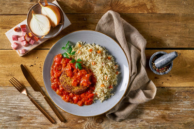 Ein Teller Reis mit Gemüse und einem mit Tomaten belegten Hackbraten steht elegant da, umgeben von Besteck, Gewürzen und einem Leinentuch – wahrlich eine Überraschungsbox von HOFMANNs, die Restaurantqualität würdig ist.