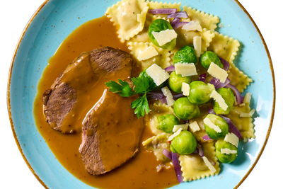 Hofmanns Shop Rinderbraten mit Hirschravioli, Rosenkohl, Zwiebeln & Parmesan auf blauem Teller.