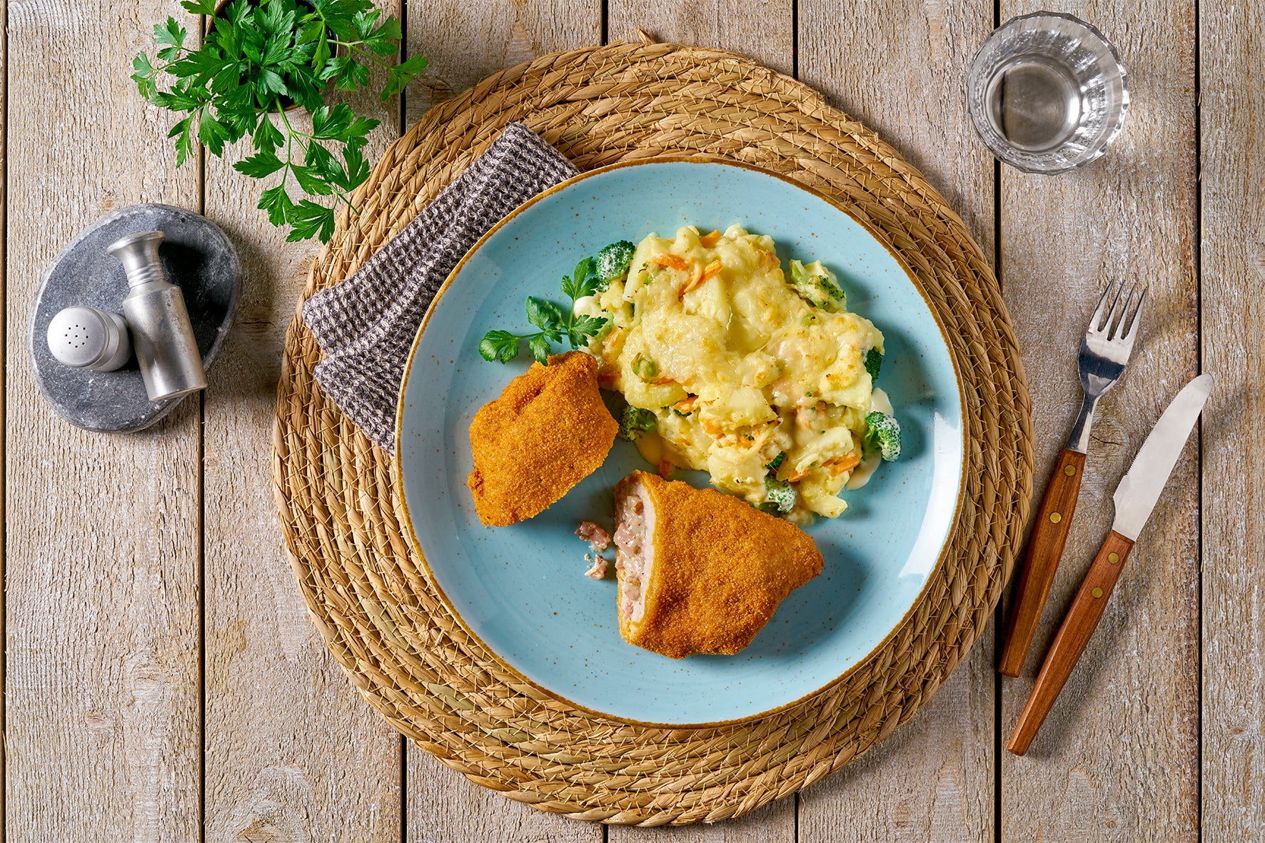 Schnitzelteller „Elsässer Art“ von Hofmanns Shop mit Käse-Brokkoli und hausgemachtem Kartoffel-Gemüse-Gratin auf gewebtem Platzset mit Besteck auf dem Tisch.