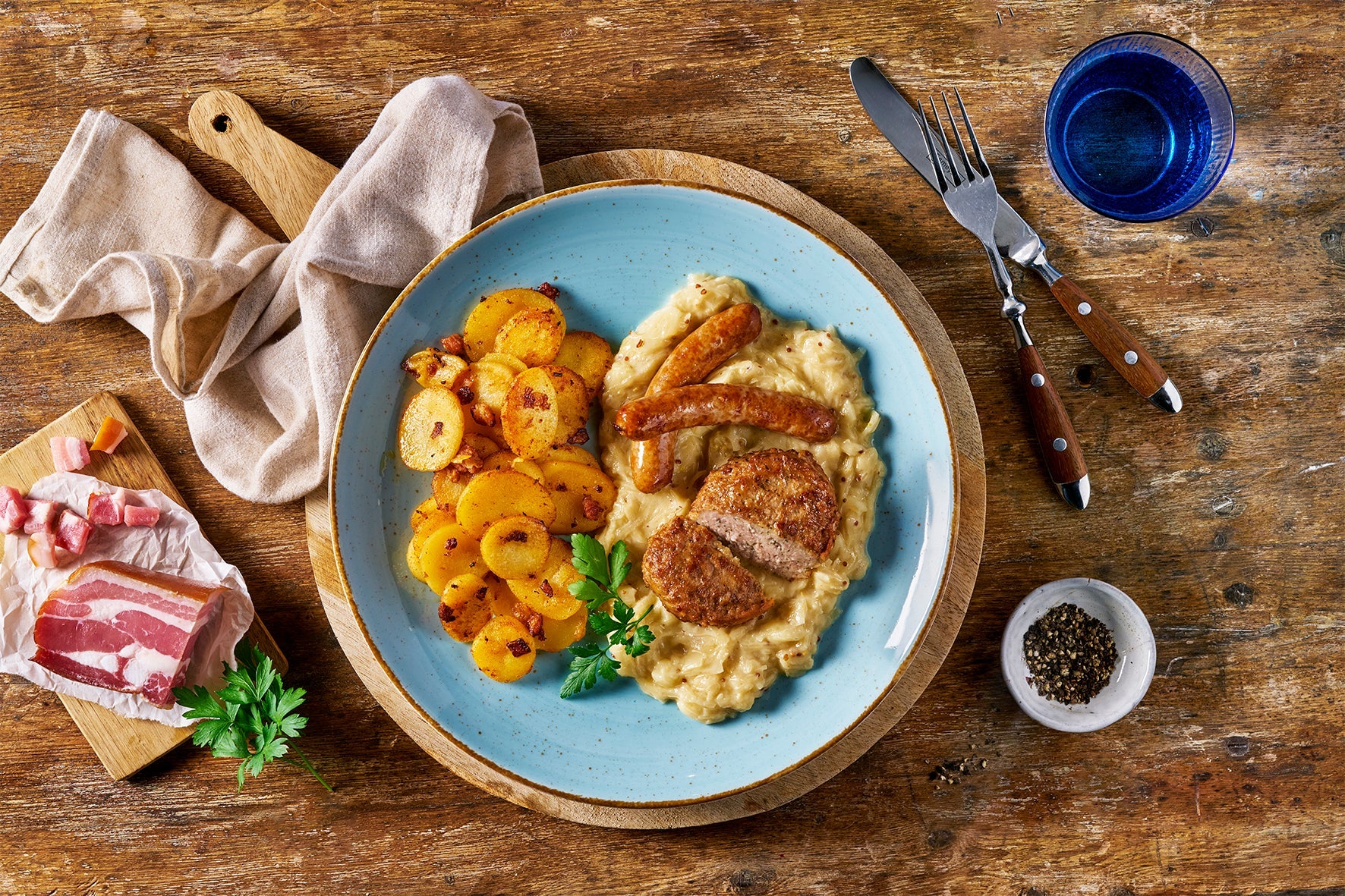 Ein blauer Teller mit Fleisch, Sahnesauce und Bratkartoffeln; Besteck und Zutaten auf einem rustikalen Holztisch präsentiert HOFMANNs Überraschungsbox in echter Restaurantqualität.