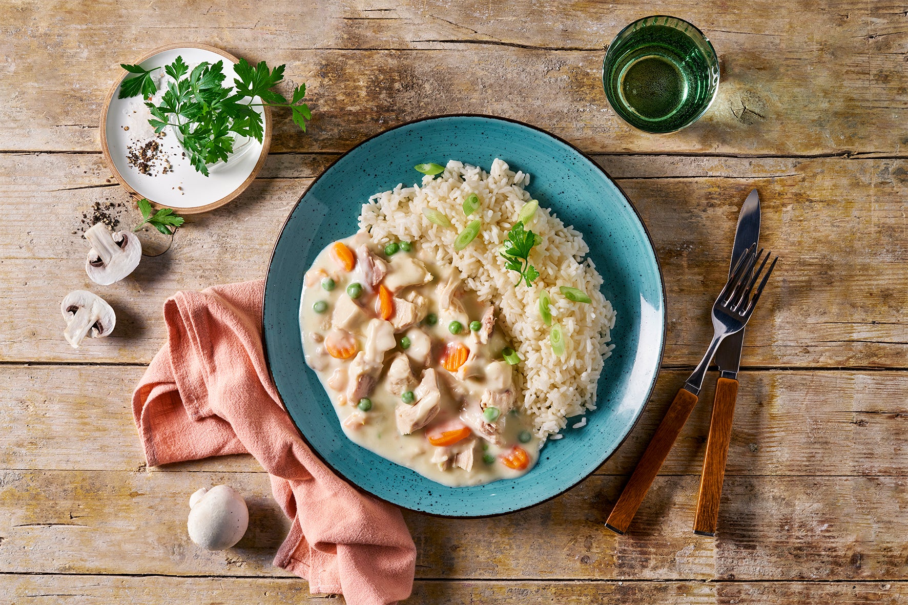 Ein Teller mit Hofmanns Shop Hühnerfrikassee und cremigem Gemüseeintopf mit Karotten, Erbsen und Champignons, garniert mit Kräutern, auf einem rustikalen Holztisch.