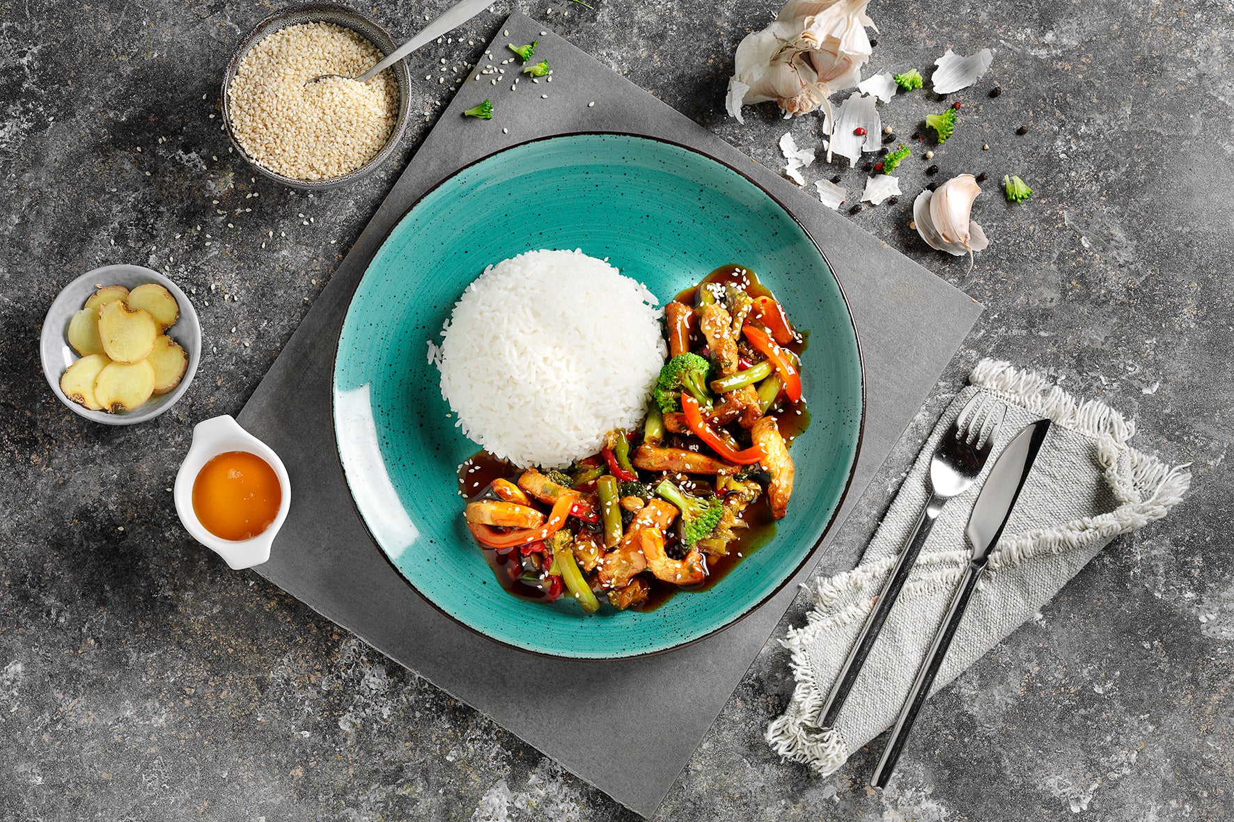 Ein blaugrüner Teller mit Basmati-Reis, gebratenem Gemüse, Hähnchen „Teriyaki Art“ von Hofmanns Shop, einer Schale Sesam, Ingwerscheiben und Besteck dazu.