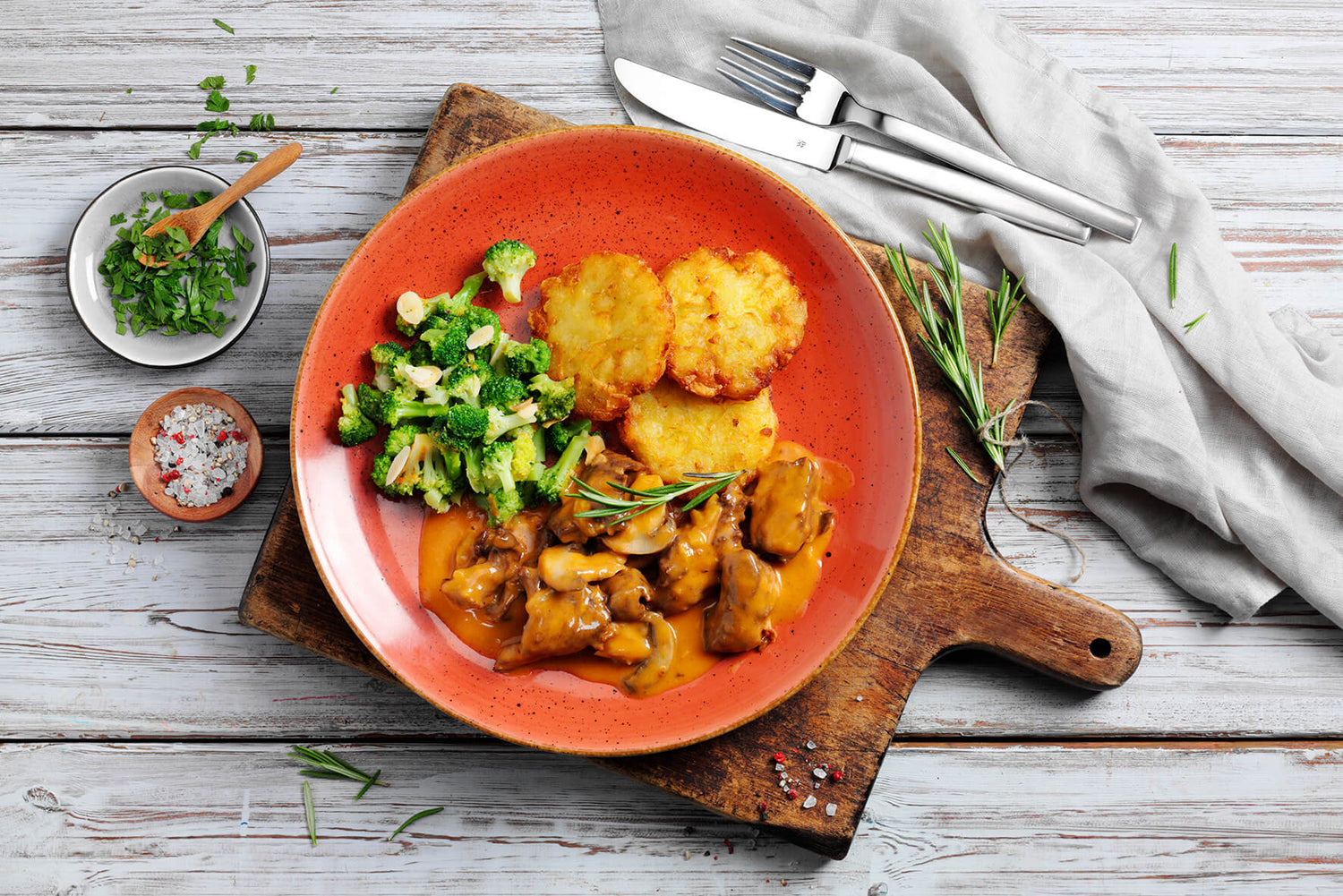 Eine HOFMANNs Festtags-Box von Hofmanns mit Bratkartoffeln, Gemüse und einem herzhaften Gericht in Soße auf einem Holzbrett mit Besteck liefert pure Genussmomente in echter Restaurantqualität.