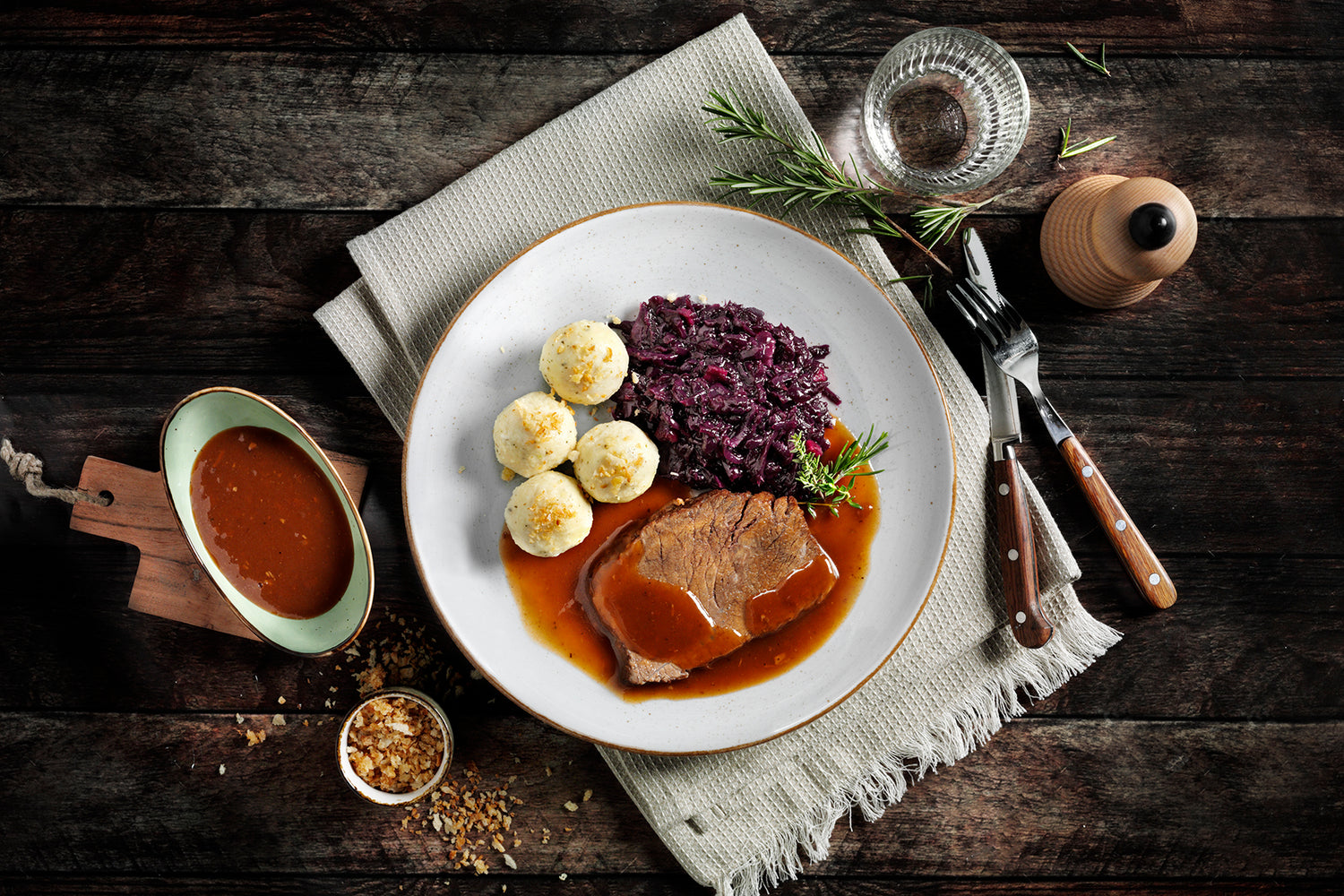 Teller mit Roastbeef, Rotkohl, Kartoffelknödeln und Soße auf einem dunklen Holztisch mit Besteck.