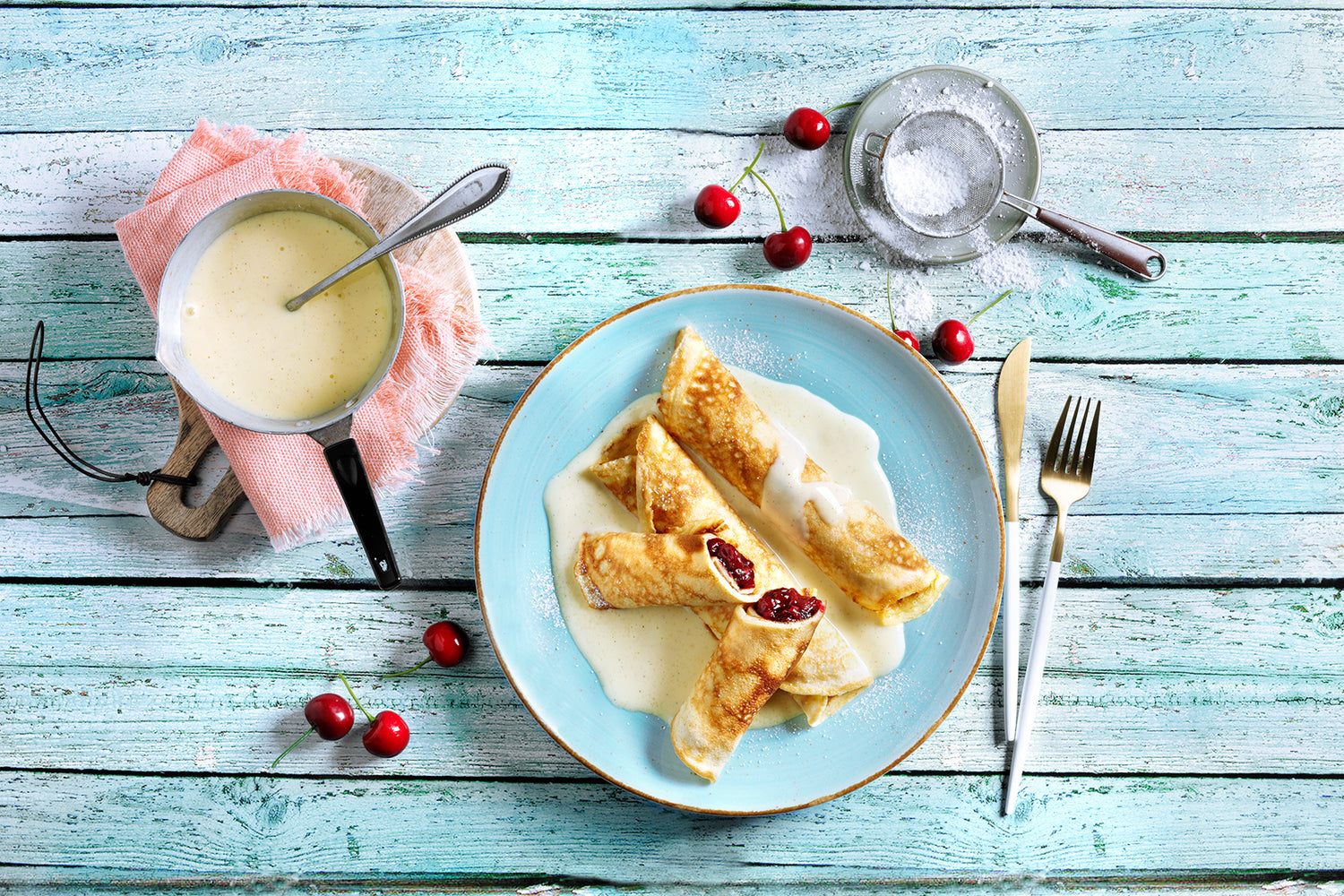 Ein Teller Crêpes mit Soße und Kirschen auf einem rustikalen Tisch, daneben ein Topf mit Soße, Gabel, Messer und frischen Kirschen.