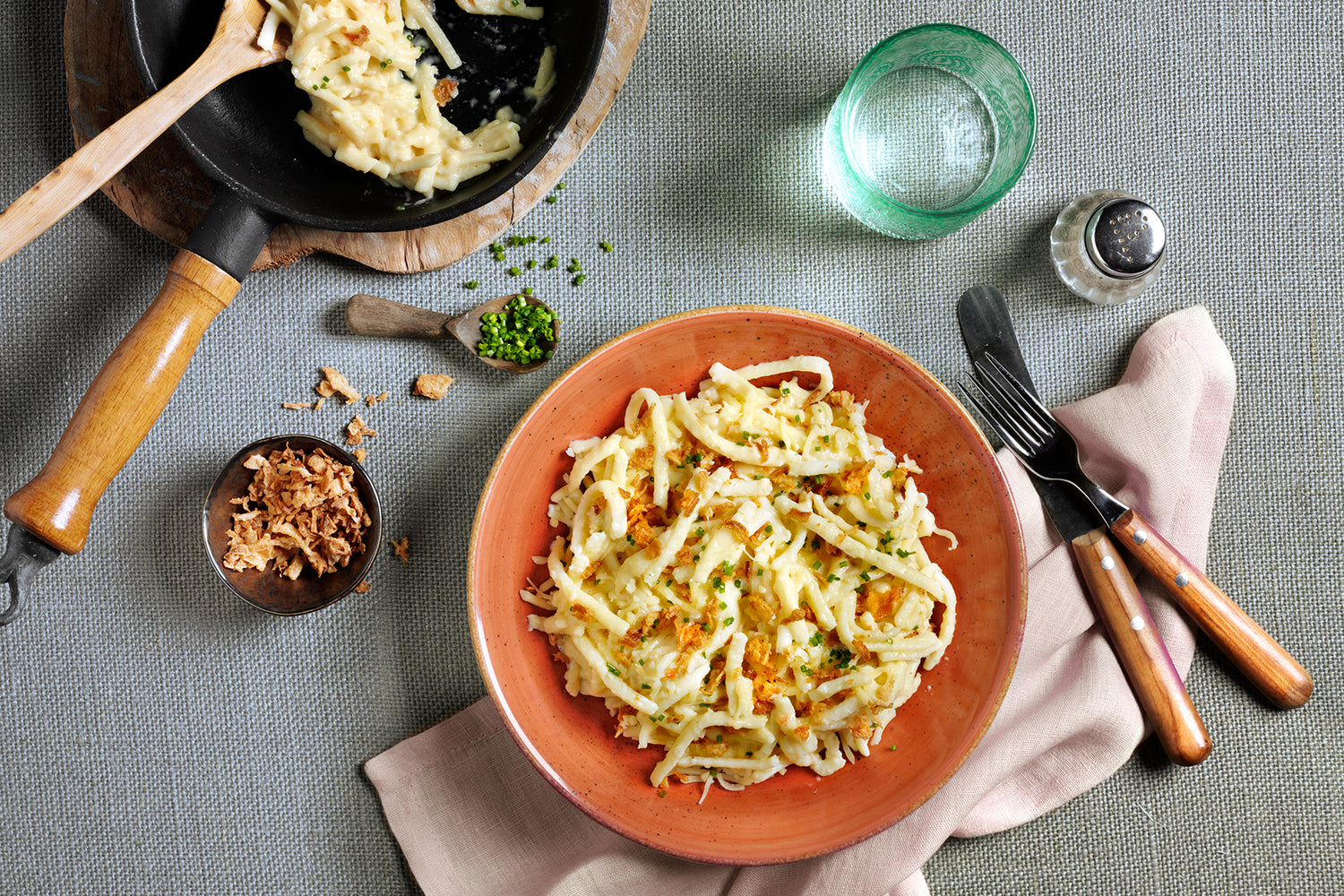 Ein Teller Käsespätzle, garniert mit Schnittlauch, steht neben einer Bratpfanne, einem Glas und Besteck auf einem Tuch und bringt den authentischen Geschmack von Omas Lieblingsrezepten aus Hofmanns Shop auf Ihren Tisch.