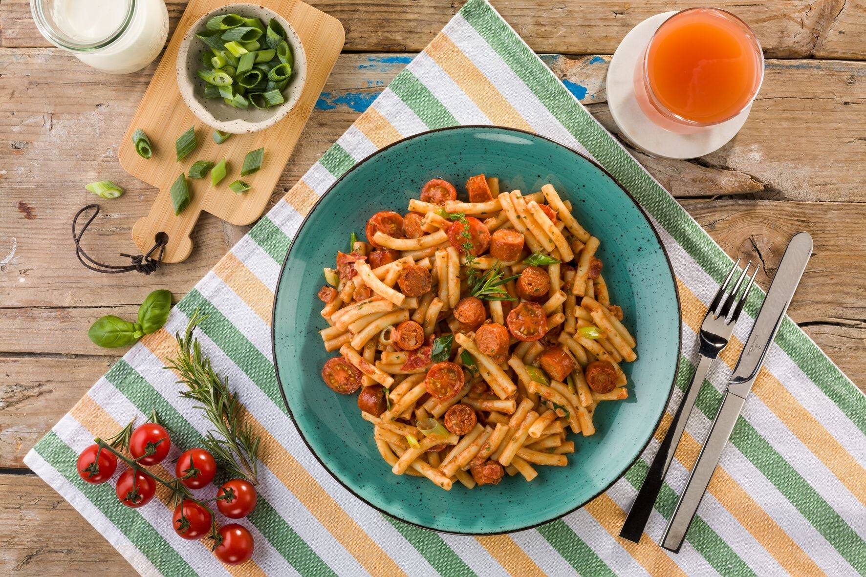 Ein Teller mit Makkaroni „Chorizo Style“ von Hofmanns Shop mit Würstchen, Kräutern und Tomaten in einer reichhaltigen Tomaten-Rahmsoße steht neben einem Glas Saft, geschnittenen Frühlingszwiebeln und einem Joghurt. Dieses Premium-Tiefkühlmenü bietet ein exquisites kulinarisches Erlebnis.