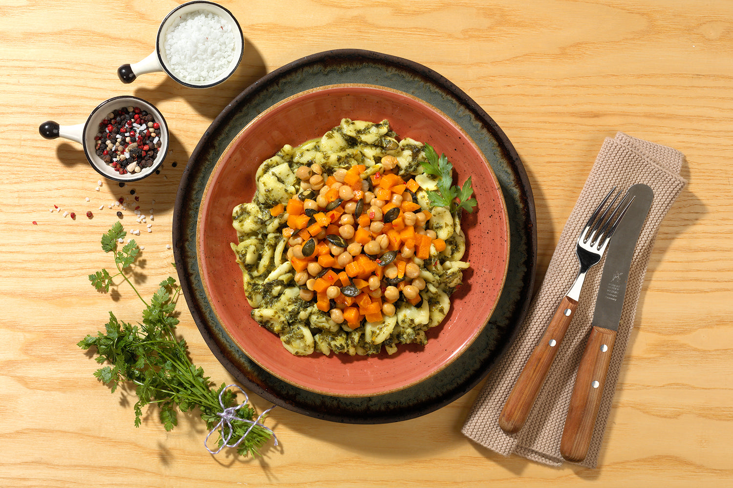 Ein Teller mit Grünkohl-Kichererbsen-Nudeln von Hofmanns Shop mit Pesto, Kichererbsen und gehackten Karotten neben Gewürzschüsseln und Utensilien auf einem Tisch, auf dem köstliche vegetarische Zutaten präsentiert werden.