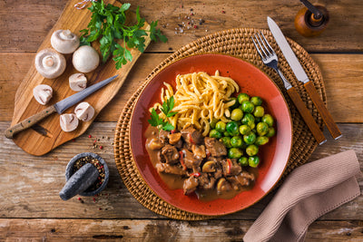 Eine Portion Hirschpfeffer-Eintopf von Hofmanns Shop, dazu Rosenkohl und Eierspätzle, steht auf einer geflochtenen Matte, umgeben von Pilzen und Petersilie, auf einem Holztisch.