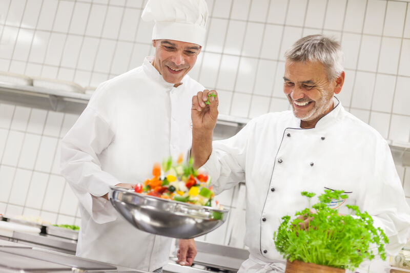 Zwei lächelnde Köche in weißen Uniformen bereiten in einer Küche mit gefliesten Wänden einen Salat zu.