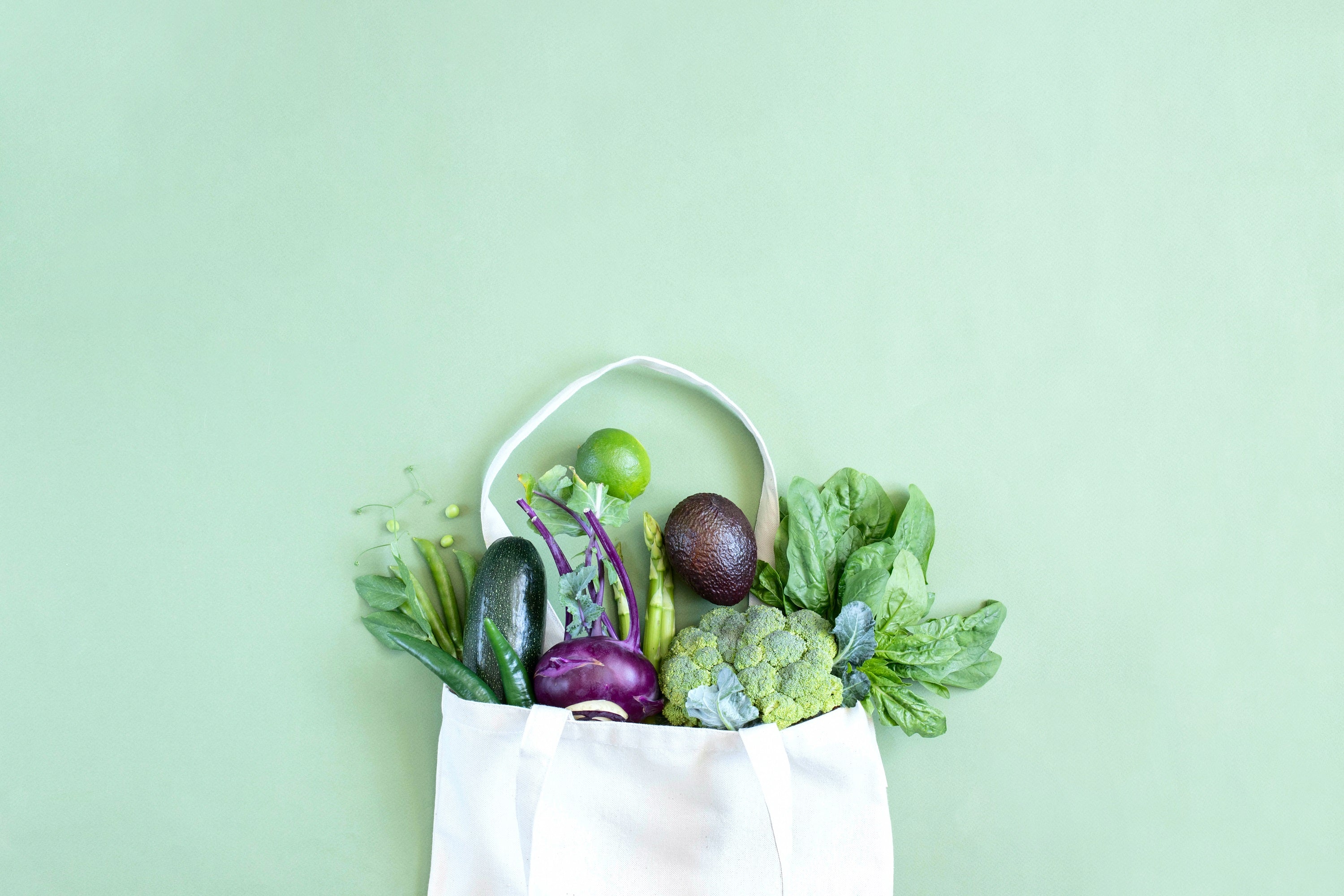 Eine Einkaufstasche gefüllt mit verschiedenen Gemüsesorten, darunter Brokkoli, Gurken und Avocado vor einem grünen Hintergrund.