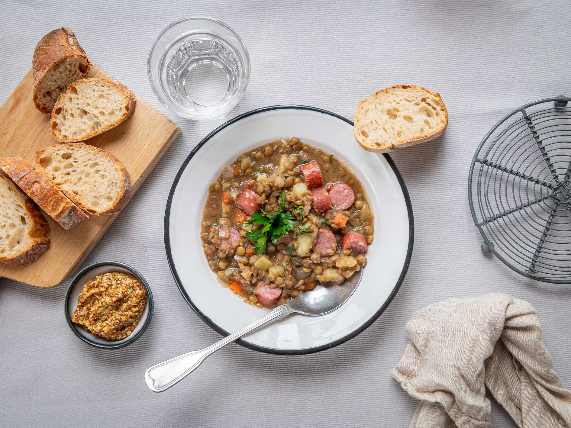 Gönnen Sie sich eine herzhafte Schüssel Linseneintopf von Hofmanns Shop mit herzhafter Wurst und Mettwurst, serviert mit geschnittenem Brot, würzigem Senf und einem erfrischenden Glas Wasser.