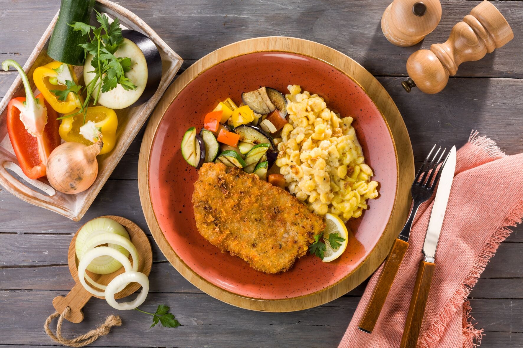 Ein Teller mit Knusperschnitzel, Rührei und geröstetem Gemüse von Hofmanns Shop, umgeben von verschiedenen Zutaten und Utensilien.