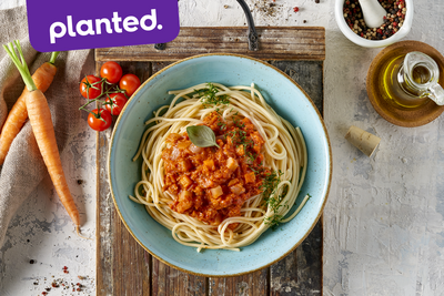 Schüssel Planted Bolognese von Hofmanns Shop mit Tomatensauce auf einem Holzbrett, umgeben von frischem Gemüse, Öl und Gewürzen.