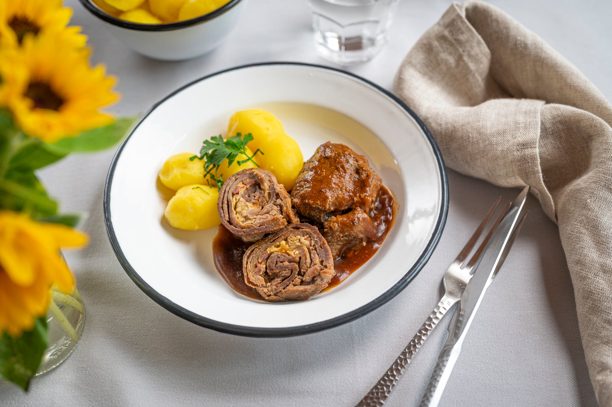 Teller mit HOFMANNs Rinderroulade aus Hofmanns Shop, mit Soße beträufelt, dazu Salzkartoffeln und eine Prise Petersilie, daneben leuchtende Sonnenblumen, eine Gabel, ein Messer und eine Serviette.