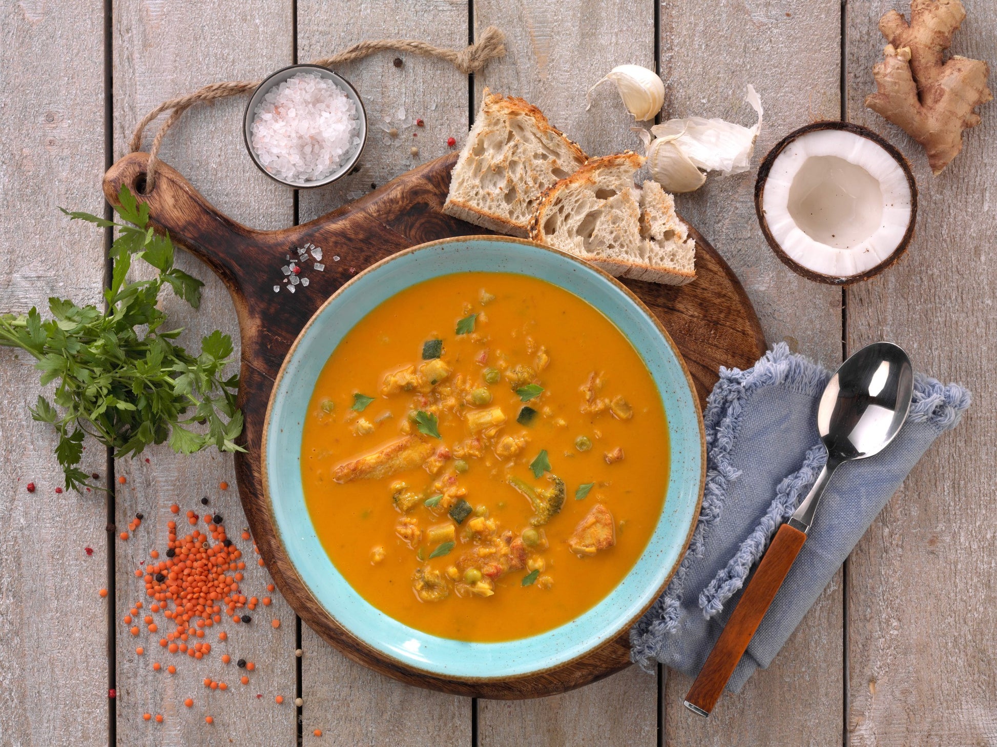 Eine Schüssel Hofmanns Rote-Linsen-Suppe auf einem Holzbrett, dazu Brot, Salz, Petersilie, Knoblauch und ein Löffel auf einer Serviette – perfekt für alle, die eine exotische Zubereitungsart schätzen.