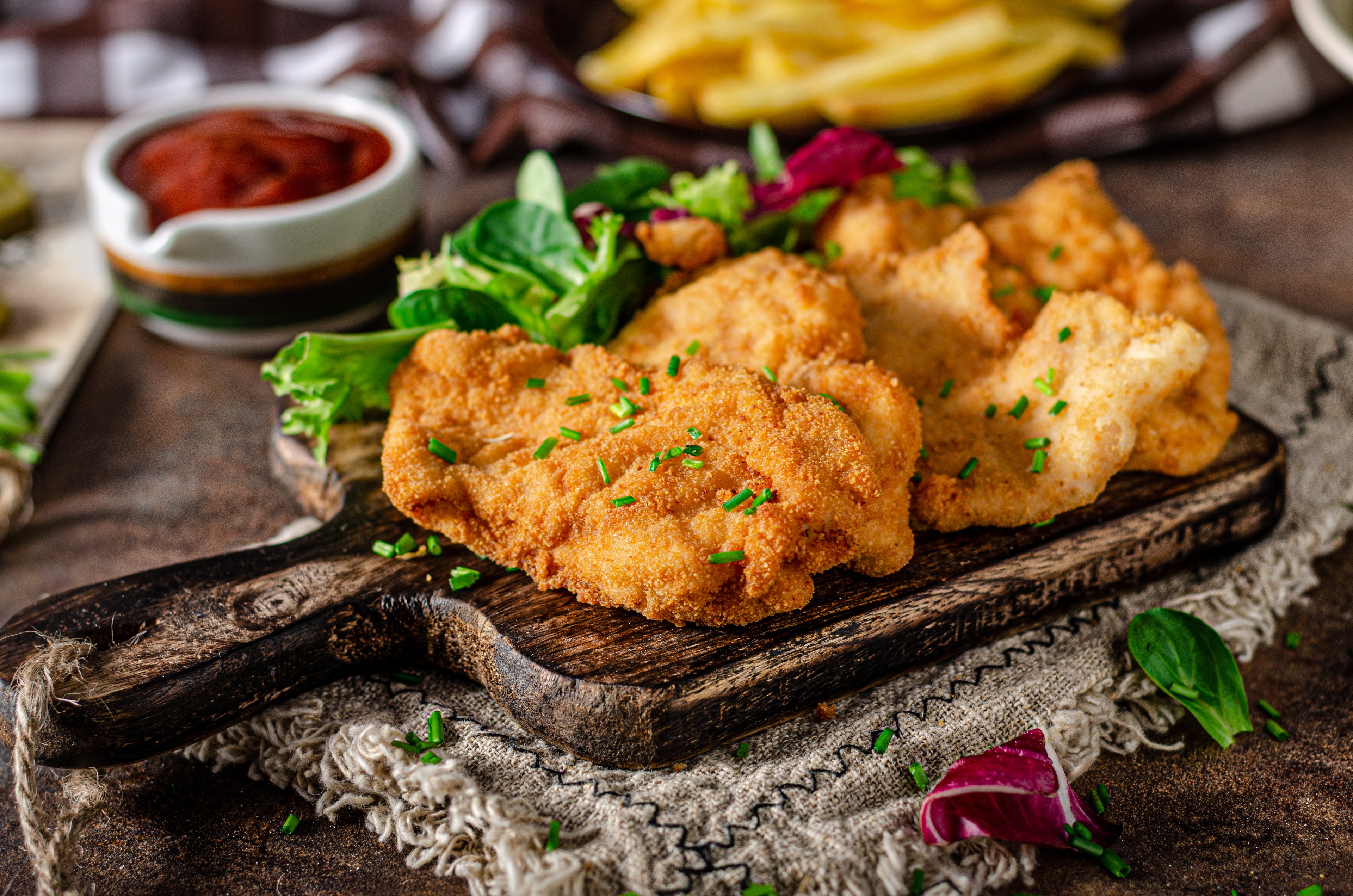 Panierte Hähnchenschnitzel auf einem rustikalen Holzbrett, garniert mit Kräutern, dazu Gemüse und Pommes Frites.