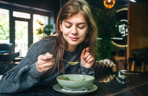 Eine Frau genießt in einem gemütlichen Café eine Schüssel Suppe und hält einen Löffel nah an ihren Mund, im Hintergrund ist Grün zu sehen.