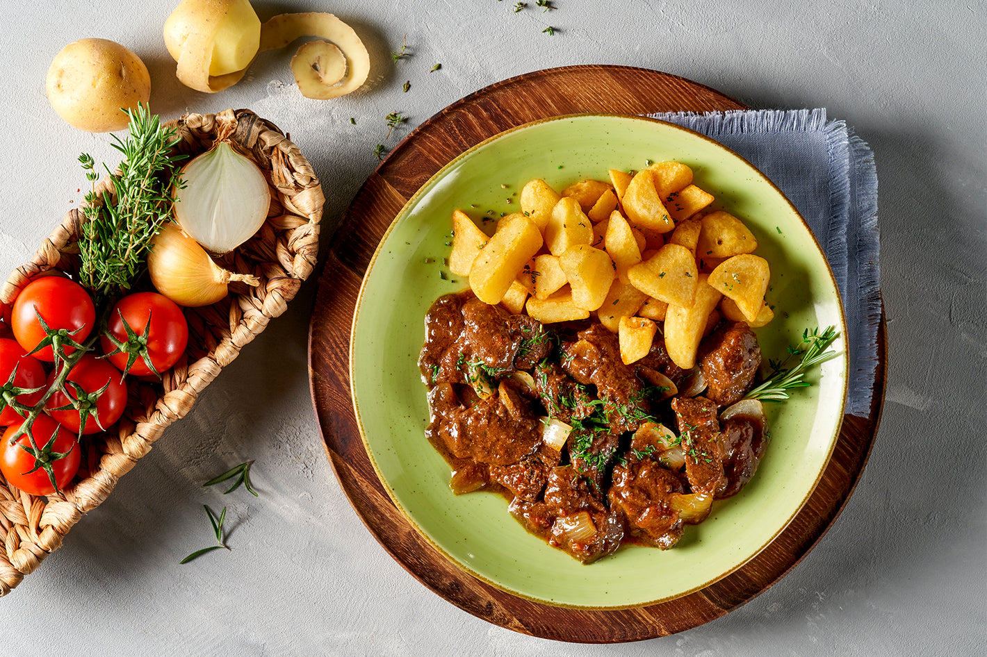 Teller mit Rindereintopf mit Kartoffeln, garniert mit Kräutern, daneben ein Korb mit Tomaten, Zwiebeln und Kartoffeln.