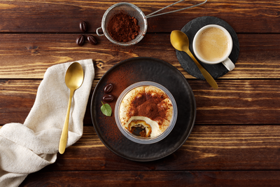 Tiramisu Klassik von Hofmanns Shop: Biskuit & Mascarponecreme auf dunklem Teller mit Kaffee & Kakaopulver.