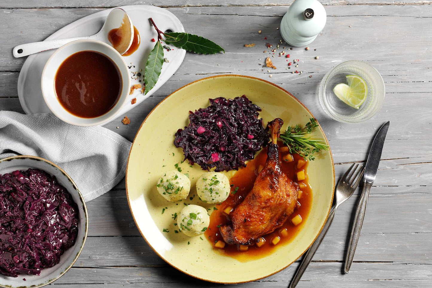 Ein klassisches Weihnachtsgericht aus der HOFMANNs Weihnachts-Box von Hofmanns Shop besteht aus einem Teller mit Entenkeule, Soße, Kartoffelknödeln und Rotkohl. Ein Beilagenteller mit Rotkohl und eine Sauciere runden dieses festliche Essen für die Feiertage ab.