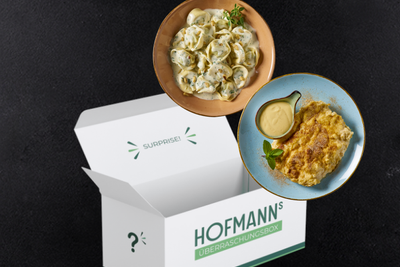 HOFMANNs Überraschungsbox mit köstlichen Knödeln und einem Käsegericht, elegant auf Tellern vor schwarzem Hintergrund präsentiert, verspricht Köstlichkeiten in Restaurantqualität, die perfekt für jeden Gourmet-Anlass sind.