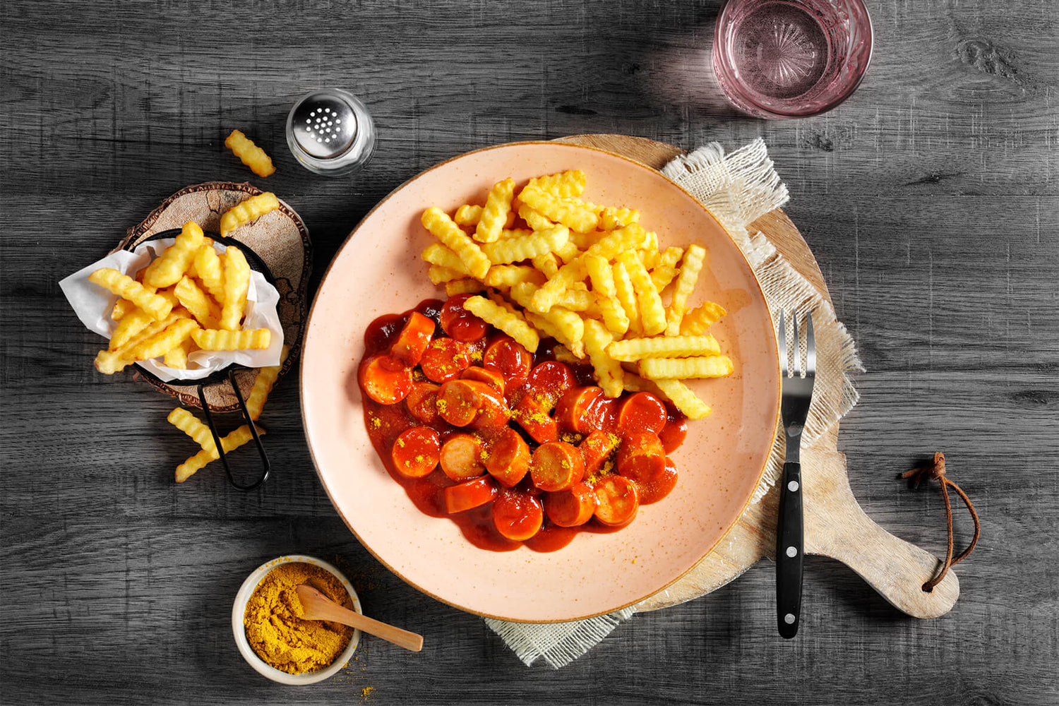 Genießen Sie einen köstlichen Teller Currywurst und knusprige Pommes, großzügig mit Currypulver bestreut, serviert auf einem rustikalen Holztisch – ideal zum Teilen. Ergänzen Sie Ihre Mahlzeit mit einem erfrischenden Glas Wasser, während Sie die Hofmanns Shop HOFMANNs Fan-Box genießen, perfekt zum Feiern klassischer deutscher Spezialitäten.