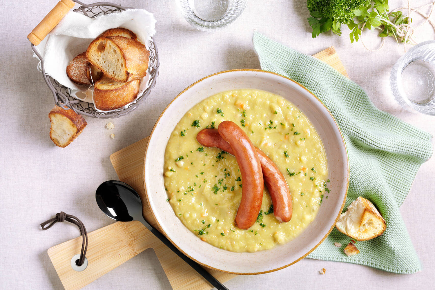 Eine Schüssel Hofmanns Erbseneintopf mit Würstchen, garniert mit Petersilie, dazu eine Scheibe Brot und ein Korb mit Brotscheiben als Beilage, sorgt für den herzhaften Geschmack der gutbürgerlichen Küche.