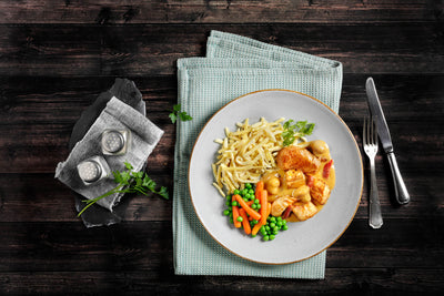Die HOFMANNs Osterbox von Hofmanns Shop besteht aus Hähnchen mit Nudeln, Erbsen und Karotten auf einer grünen Unterlage und strahlt Restaurantqualität aus. Besteck und Gewürzstreuer daneben runden das Festtagsgericht-Ambiente ab.