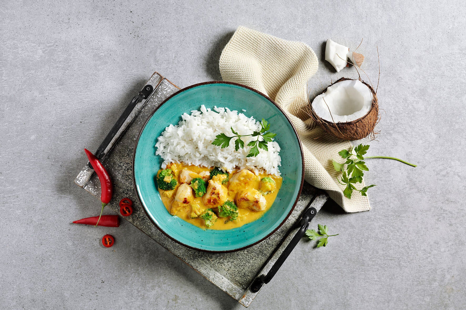 Eine Feinschmecker-Box von Hofmanns mit einer blauen Schale gefüllt mit Hühnercurry, Brokkoli und weißem Reis. Garniert mit Petersilie für pure Genussmomente, dazu rote Chilis und Kokosnuss als Beilage.