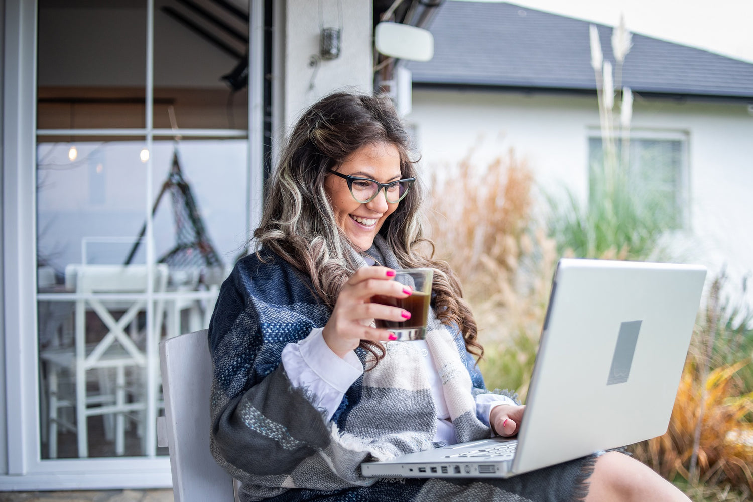 Frau sitzt auf der Terrasse mit einem Laptop und Kaffee in der Hand.