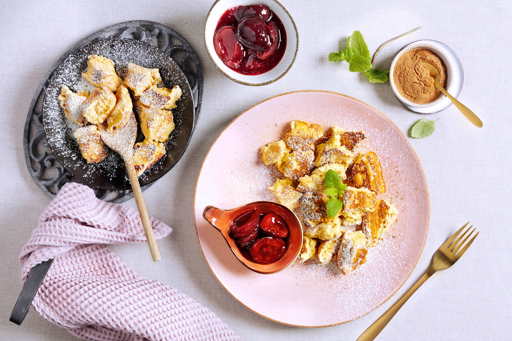 Zwei Teller Kaiserschmarrn mit Puderzucker, Pflaumenkompott, Zimt und einer rosa Serviette auf einem weißen Tisch.