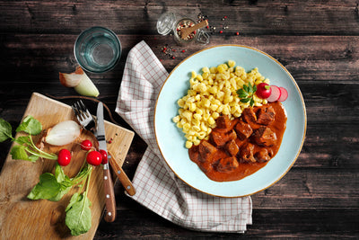 Genießen Sie eine Portion Gulasch und Nudeln neben einem Schneidebrett mit Radieschen, Zwiebeln, Gabel, Messer und einem erfrischenden Glas Wasser – ein authentischer Hochgenuss in Restaurantqualität aus HOFMANNs Osterbox von Hofmanns Shop.