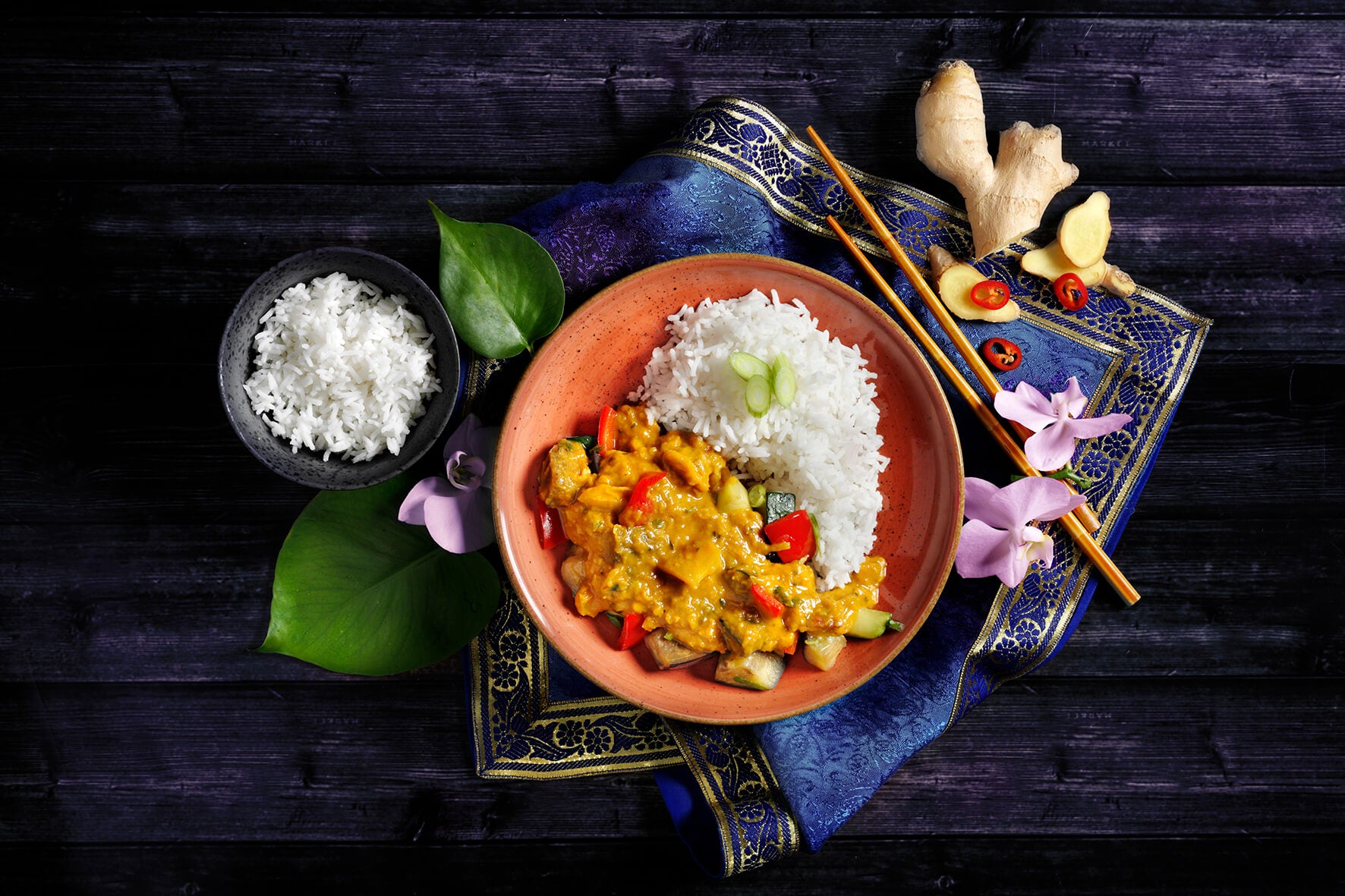 Ein Gericht aus Reis-Gemüse-Curry, garniert mit Frühlingszwiebeln, begleitet von Ingwer und Orchideenblüten, wird in der Vegan-Box von Hofmanns Shop zu einem unschlagbaren Preis angeboten.