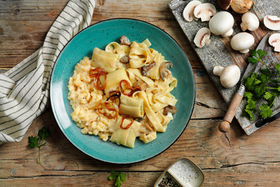 Ein Teller Pappardelle-Nudeln mit Pilzen, Zwiebeln und einer cremigen Sauce steht neben einem Tuch und einem Schneidebrett und präsentiert eines unserer vegetarischen Bestseller-Gerichte aus der Probierbox: Fleischfreie Alternativen von Hofmanns Shop.