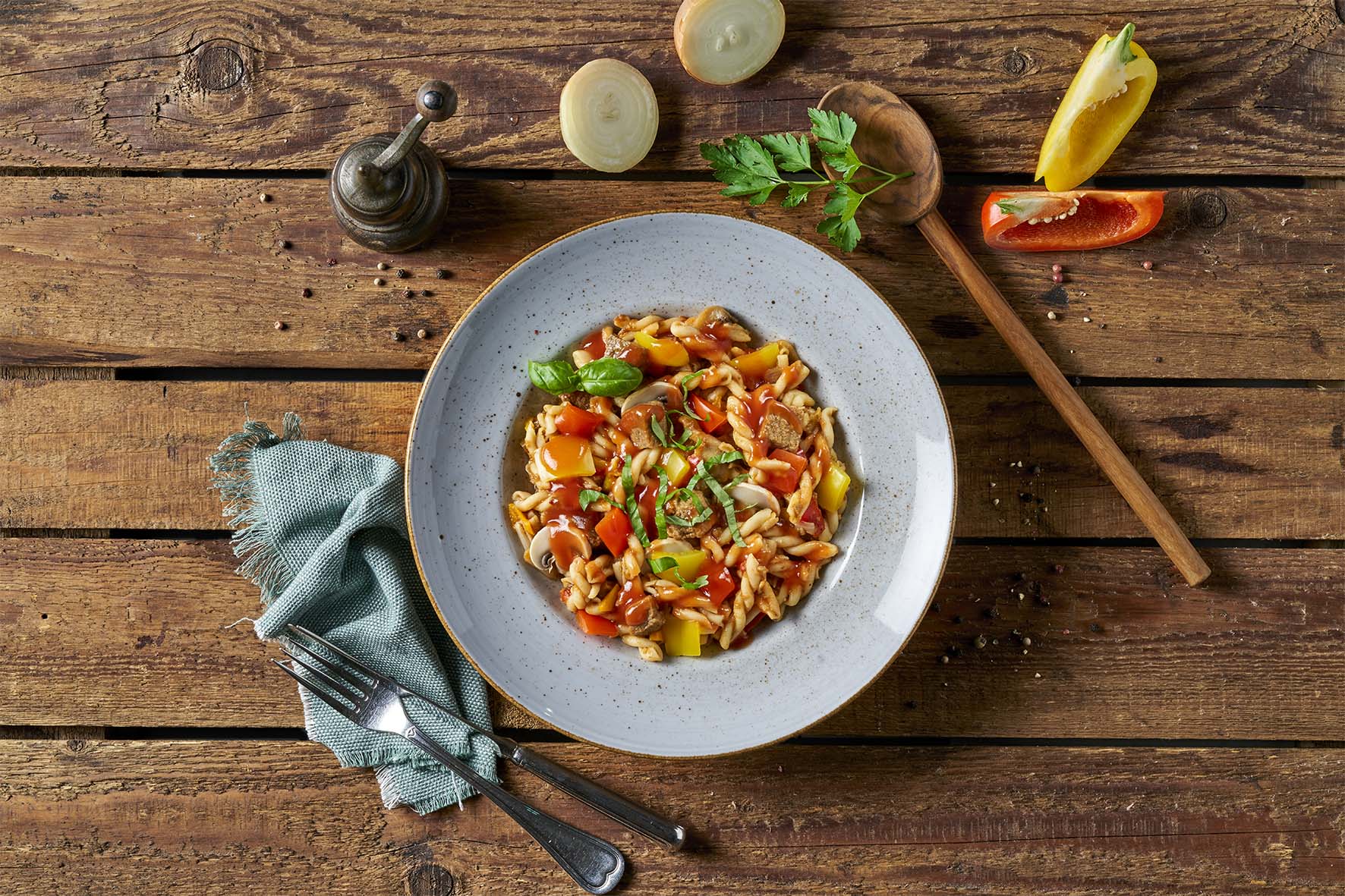 Eine Schüssel Pasta mit Gemüse auf einem rustikalen Holztisch, begleitet von Besteck und Gewürzen, spiegelt die Qualität der HOFMANNs Überraschungsbox wider.