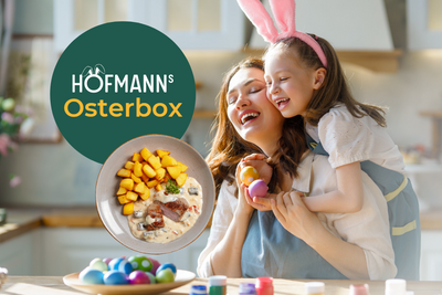 Eine Frau und ein Kind mit Hasenohren lächeln in einer Küche mit der HOFMANNs Osterbox von Hofmanns Shop, gefüllt mit festlichem Geschirr und dekorierten Eiern.
