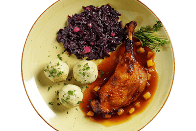 Ein Teller mit Hofmanns Entenkeule, serviert mit Knödeln, Apfelrotkohl und Rosmaringarnitur, begleitet von einer würzigen Bratapfelsoße.