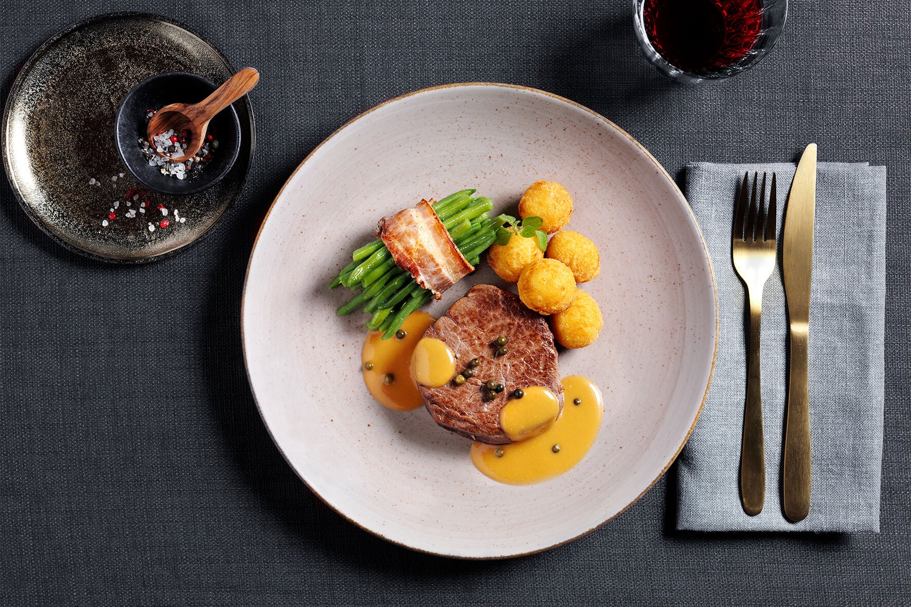 Eine Gourmet-Mahlzeit **Protein Deluxe-Box** von Hofmanns Shop auf einem weißen Teller mit Steak, grünen Bohnen, Kartoffelkroketten und würziger Sauce, perfekt kombiniert mit einem Glas Rotwein.