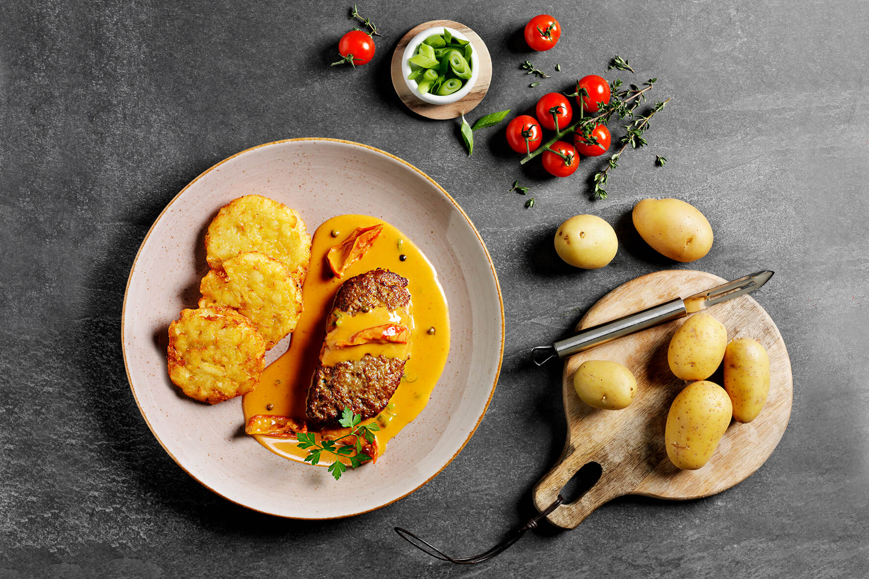 Teller mit Hacksteak „Gipfelstürmer“, Sauce und Rösti, dazu Kartoffeln, Kirschtomaten, Kräuter und Frühlingszwiebeln – ein Hofmanns Tiefkühlmenü, das ein Premiumgericht-Erlebnis bietet.