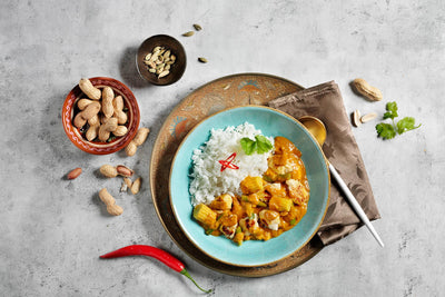 Hähnchen Erdnuss-Tikka von Hofmanns Shop mit Reis, Chili & Kräutern auf Tablett, grauer Untergrund.