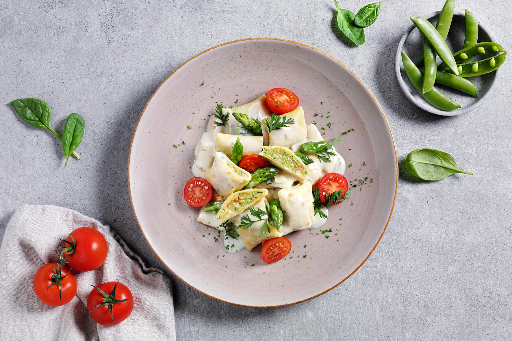 Ein Teller Ravioli mit Kirschtomaten, Spargel und Kräutern auf grauem Untergrund erinnert an Omas Verwöhnmenü aus Hofmanns Shop und erweckt herzhafte und klassische Aromen zum Leben.