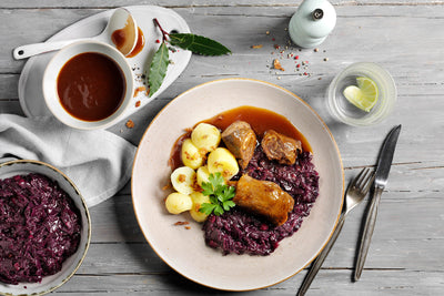 Teller mit Rinderrouladen, Rotkohl und Kartoffeln auf einem Holztisch, dazu Bratensoße und Wasser. Diese HOFMANNs Osterbox aus Hofmanns Shop bietet Hochgenuss in Restaurantqualität.