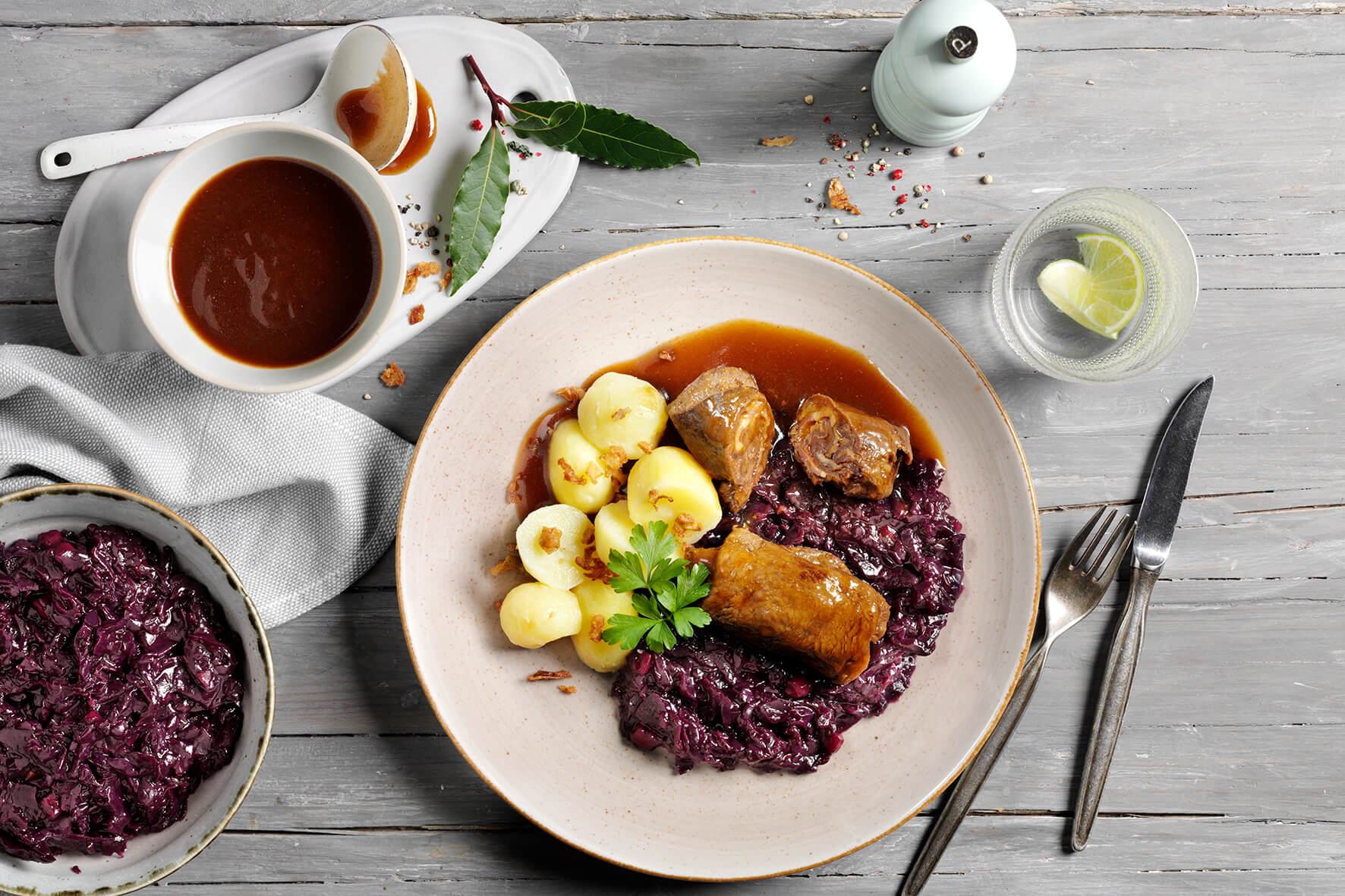 Teller mit Rinderrouladen, Rotkohl und Kartoffeln auf einem Holztisch, dazu Bratensoße und Wasser. Diese HOFMANNs Osterbox aus Hofmanns Shop bietet Hochgenuss in Restaurantqualität.